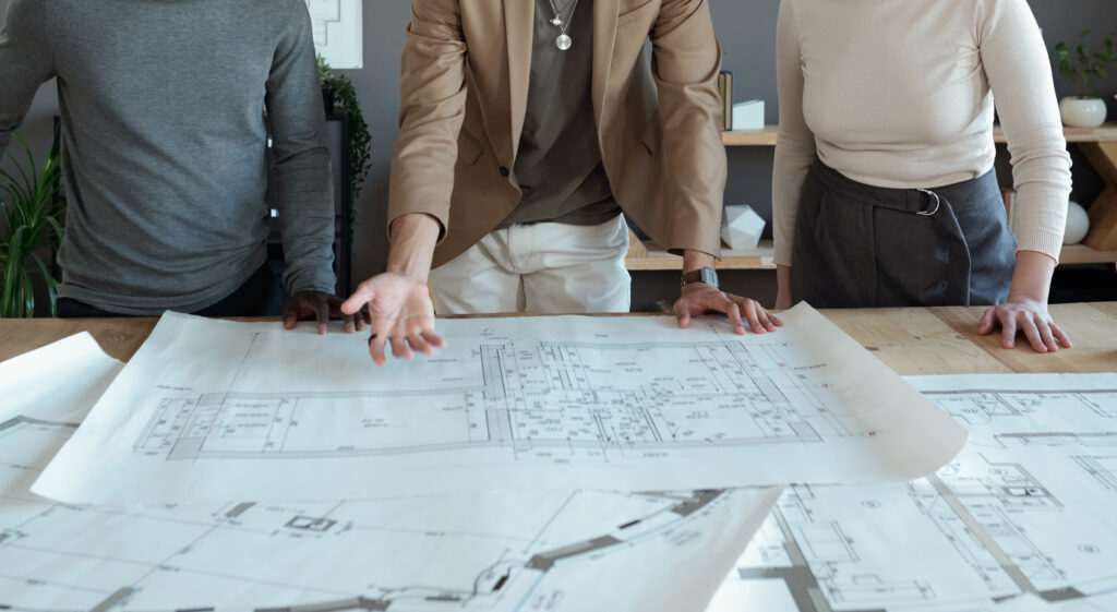 One of three coworkers making presentation of large sketch on blueprint and explaining it to intercultural colleagues