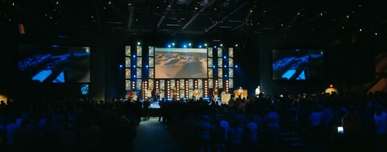 Congregation facing a lit stage and screens beneath an exposed auditorium ceiling